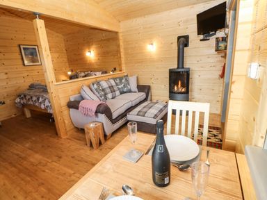 A wooden cabin interior with a dining table set for two a sofa with cushions a wood stove and a bed in a separate area at Otters Holt in Winston near Staindrop