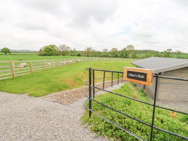 A gravel driveway with a black metal gate and a sign reading Otters Holt a wooden building and fenced green fields with sheep at Otters Holt in Winston near Staindrop