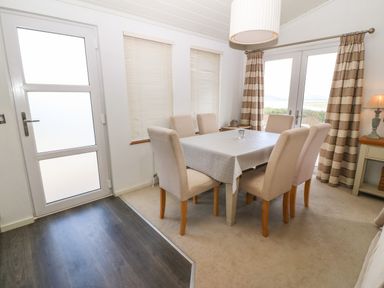 A dining room with a table covered by a tablecloth and six chairs near windows and glass doors at Sunstone Lodge Garreg Haul Lodge in Cilan near Abersoch