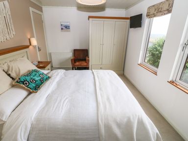 A bedroom with a bed a white wardrobe a brown chair and windows with a view at Sunstone Lodge Garreg Haul Lodge in Cilan near Abersoch