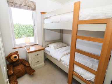 A small bedroom with bunk beds a bedside table with a lamp a window and a large teddy bear on the floor at Sunstone Lodge (Garreg Haul Lodge) in Cilan near Abersoch