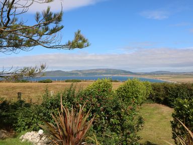 A garden with bushes and a tree overlooking a field and a body of water with hills in the background at Sunstone Lodge Garreg Haul Lodge Cilan near Abersoch