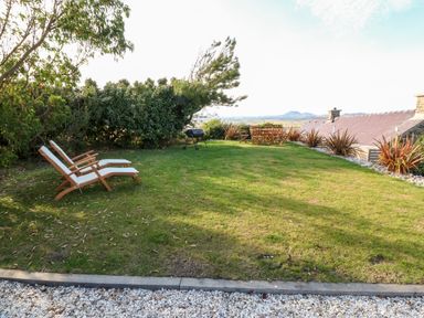 A garden with two wooden lounge chairs a barbecue and a wooden dining table with chairs at Sunstone Lodge Garreg Haul Lodge Cilan near Abersoch