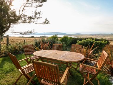 A wooden outdoor table with six wooden chairs on grass with bushes and a scenic view of fields and hills at Sunstone Lodge Cilan near Abersoch