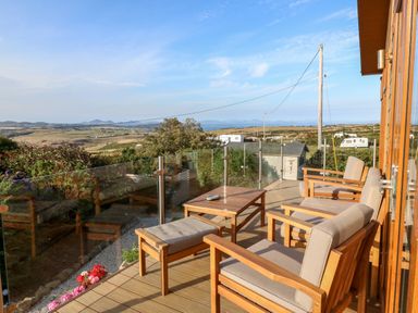 An outdoor patio with wooden chairs and a table overlooking a rural landscape with hills and caravans at Sunstone Lodge Garreg Haul Lodge in Cilan near Abersoch