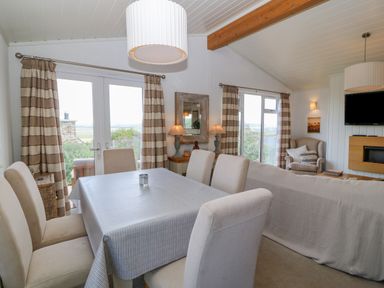 A dining area with a table and chairs next to a living room with a sofa armchair and a wall-mounted TV at Sunstone Lodge Garreg Haul Lodge in Cilan near Abersoch