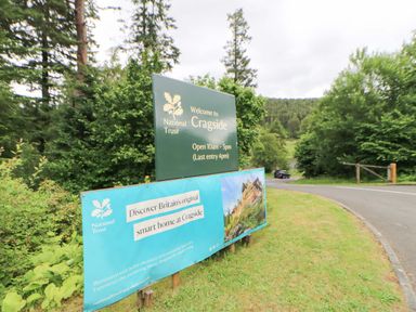 A sign for Cragside with visiting information and a car on the road at Cragside in Rothbury