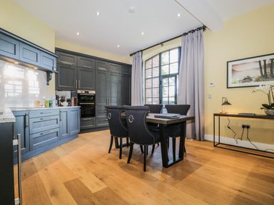 A dining area with a wooden table and black chairs beside a kitchen with dark cabinetry and a large window with curtains at Tarn End Cottages 5 Talkin near Brampton Cumbria