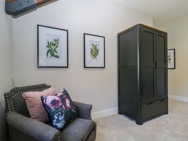 A gray armchair with floral pillows next to a dark wooden wardrobe with botanical prints on the wall at Tarn End Cottages 5 Talkin near Brampton Cumbria