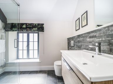 A bathroom with a window covered by a patterned blind a toilet a sink with a wooden cabinet and framed pictures above the sink at Tarn End Cottages 5 Talkin near Brampton Cumbria