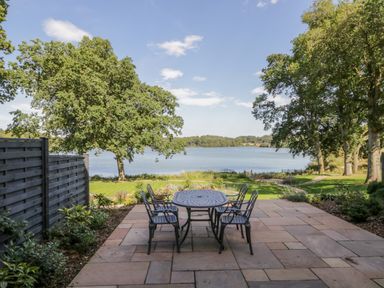 A patio with a metal table and six chairs overlooking a lawn with trees and a lake at Tarn End Cottages 5 Talkin near Brampton Cumbria