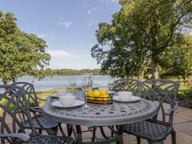 An outdoor metal dining table set with bowls plates wine glasses and a basket of lemons near a lake with trees at Tarn End Cottages 5 Talkin near Brampton Cumbria
