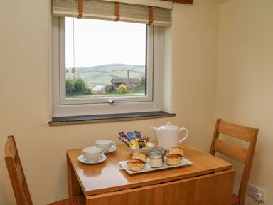 A small wooden dining table with two chairs set with cups saucers a teapot scones and a bowl of tea bags by a window with a countryside view at Beech Cottage in Combe Martin