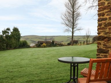 A grassy yard with a round glass table and wooden chairs next to a stone building with hills in the background at Beech Cottage in Combe Martin
