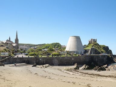 A sandy beach with rocks and stone wall overlooking buildings including a church with a tall spire and a large conical structure in a coastal town