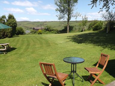 A grassy outdoor area with two wooden chairs and a round glass table with hills in the background at Beech Cottage in Combe Martin