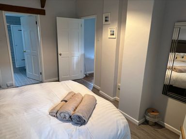 A bedroom with a bed covered in white bedding and three rolled towels on top two open doors leading to adjacent rooms and a mirror on the wall near a small trash bin