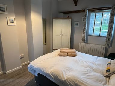 A bedroom with a bed with white linens and towels on top a white wardrobe a window with blinds and curtains and wooden floor at Beech Cottage in Combe Martin