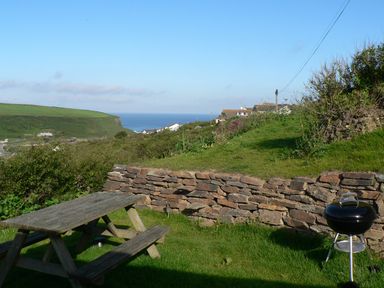 An outdoor picnic table and a barbecue grill on grass with a stone wall and houses on a hill and the ocean in the background at Porth Farm Annexe in Mawgan Porth