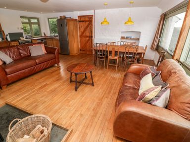 An open living room and kitchen with two leather sofas a wooden dining table and chairs and a basket of firewood at Porth Farm Annexe in Mawgan Porth