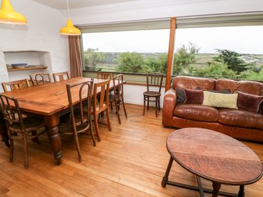 A dining area with a wooden table and chairs next to a brown leather sofa and a wooden coffee table at Porth Farm Annexe in Mawgan Porth