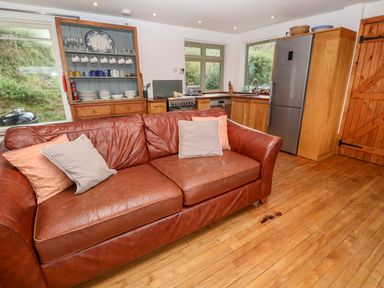 A kitchen and living area with a brown leather sofa wooden cabinets and a refrigerator at Porth Farm Annexe Mawgan Porth