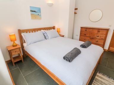 A bedroom with a wooden bed with striped bedding two bedside tables with lamps a wooden dresser with a round mirror and a painting above the bed at Porth Farm Annexe in Mawgan Porth