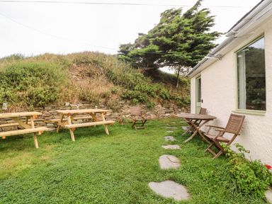 An outdoor area with wooden picnic tables and chairs beside a white building at Porth Farm Annexe in Mawgan Porth