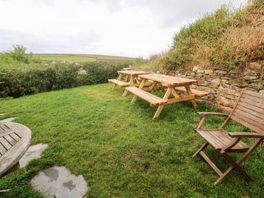 A grassy outdoor area with wooden picnic tables and a wooden folding chair at Porth Farm Annexe in Mawgan Porth
