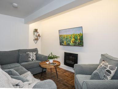 A living room with grey sofas a wall-mounted TV showing sunflowers and a round wooden coffee table at Y Goedwig in Newborough