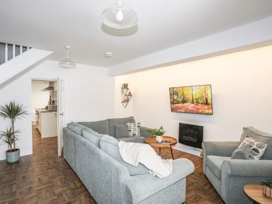 A living room with a grey sectional sofa an armchair a round wooden coffee table a wall-mounted television a potted plant and an open doorway showing a kitchen at Y Goedwig in Newborough
