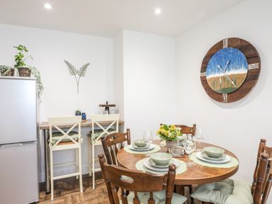 A dining area with a round wooden table set with dishes and glasses and wooden chairs with cushions at Y Goedwig in Newborough
