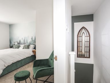 A bedroom with a bed a chair and a small stool and a hallway with a tall arched mirror at Y Goedwig in Newborough