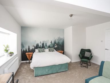 A bedroom with a bed a forest mural green armchair and plants on the windowsill at Y Goedwig in Newborough