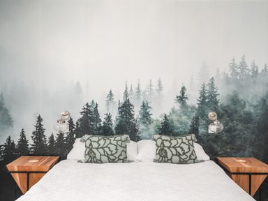 A bedroom with a bed featuring white bedding and green patterned pillows with wooden nightstands under wall-mounted glass light fixtures and a forest-themed mural behind the bed at Y Goedwig in Newborough