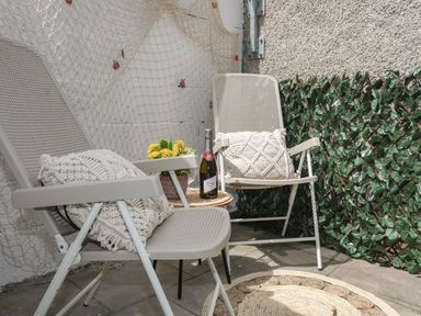 An outdoor seating area with two beige chairs having cushions a small round table with a plant and a bottle a wall with netting and decorative green foliage at Y Goedwig in Newborough