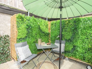 A small patio with two chairs a table an umbrella and green plants on wooden walls at Y Goedwig in Newborough