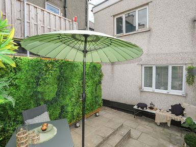 An outdoor patio with a green umbrella a small table gray chairs and a bench with cushions at Y Goedwig in Newborough