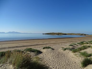A sandy beach with grassy dunes leading to calm water and a small island in the distance at Y Goedwig in Newborough