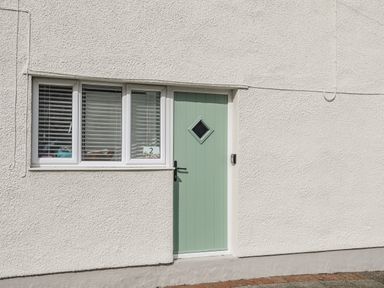 A light green door with a diamond shaped window next to three white framed windows on a white textured wall at Y Goedwig in Newborough