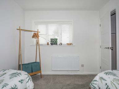 A bedroom with two beds with green leaf patterned bedding a wooden clothes rack with hangers and storage bins and a window with white blinds at Y Goedwig in Newborough