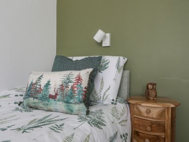 A bed with green and white patterned bedding and pillows next to a wooden bedside table with an owl figurine at Y Goedwig in Newborough