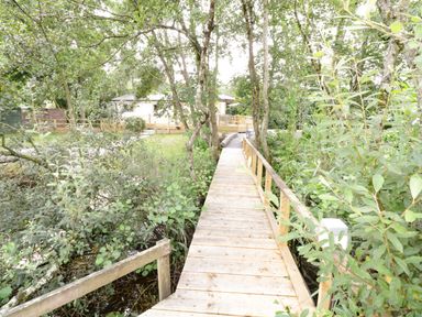 A wooden footbridge over a small stream surrounded by trees and bushes leading to a house with a deck at Port Heron Lodge Ballykeeran near Glasson County Westmeath