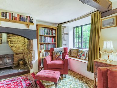 A sitting room with a red armchair and footstool a window seat with cushions a wood stove bookshelf and lamp at Saint Pancras Chapel in Washford near Williton