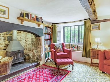 A sitting room with a stone fireplace red armchair and ottoman a window seat with cushions and bookshelves at Saint Pancras Chapel in Washford near Williton