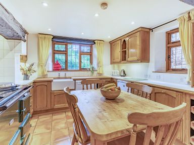 A kitchen with wooden cabinets a wooden table with chairs and a bowl of fruit by windows with yellow curtains at Saint Pancras Chapel in Washford near Williton