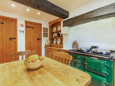 A kitchen with wooden table and chairs a bowl of fruit green stove wooden doors and shelving with plates and clock at Saint Pancras Chapel in Washford near Williton