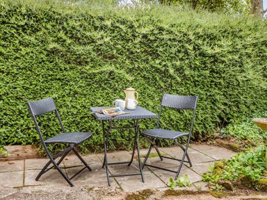 An outdoor patio with two black folding chairs and a small black table with two mugs and a jug in front of a tall hedge at Saint Pancras Chapel in Washford near Williton