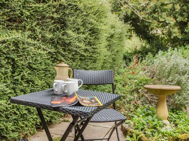 A black outdoor table with three white mugs a teapot and a magazine next to a black chair in a garden with plants and a birdbath at Saint Pancras Chapel in Washford near Williton
