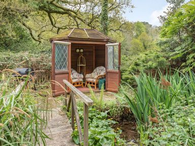 A wooden garden gazebo with open doors and cushioned chairs surrounded by plants and trees at Saint Pancras Chapel in Washford near Williton